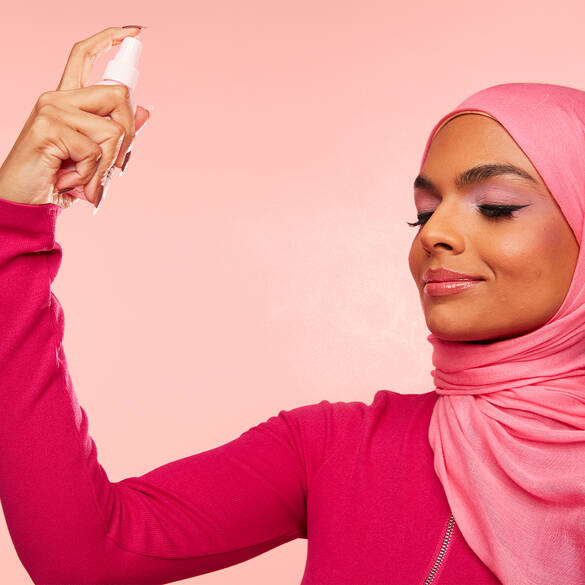 Een vrouw in een roze hijab spuit een schoonheidsproduct op haar gezicht met een glimlach, tegen een roze achtergrond.