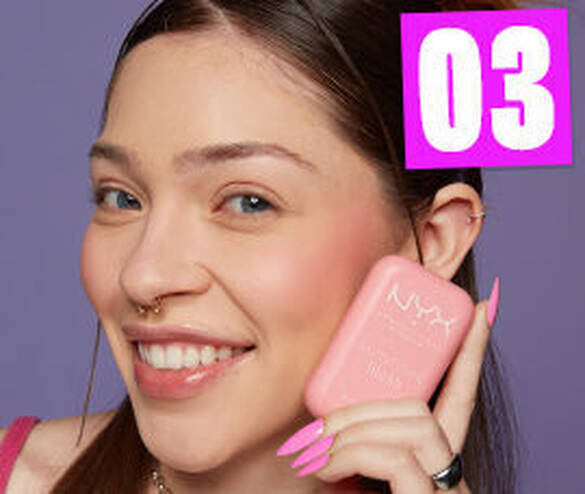 Femme souriante avec un blush NYX, tenant la boite en rose, sur fond violet. Ongles longs et roses.